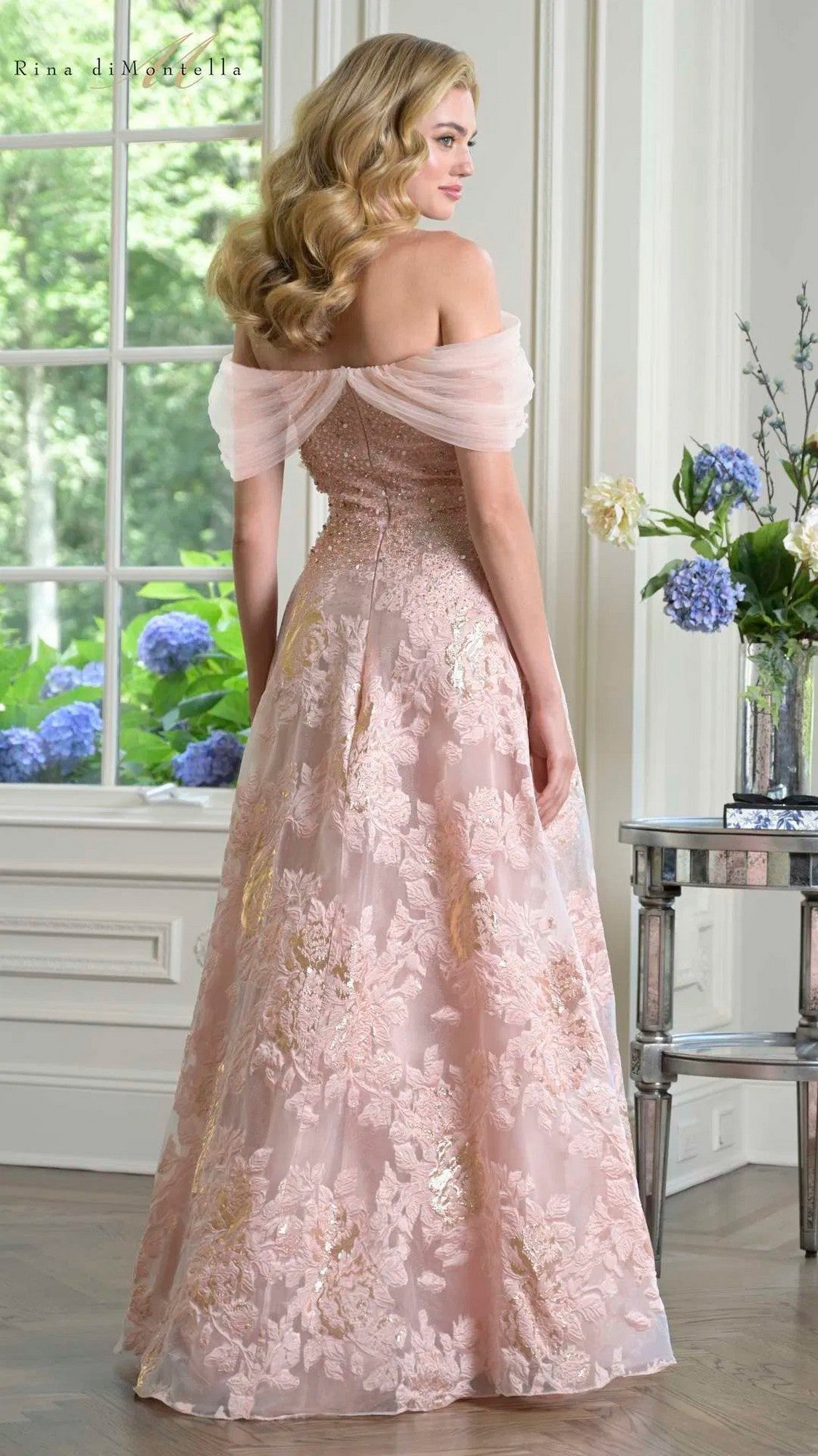 A woman stands indoors wearing the Rina Di Montella RD3406 long formal dress, which has an off-the-shoulder beaded bodice and a flowing A-line skirt. Blue hydrangeas and greenery appear through a window and in a nearby vase.